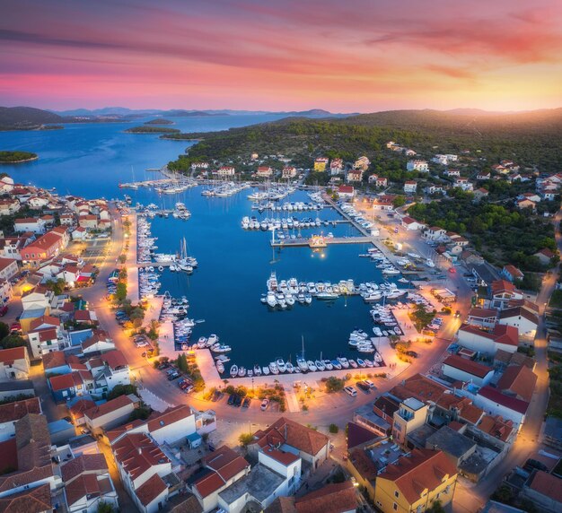 Aerial view of boats and yachts in port and city at sunset summer landscape with city lights buildings illuminated streets mountain motorboats blue sea red sky at night top view of croatia