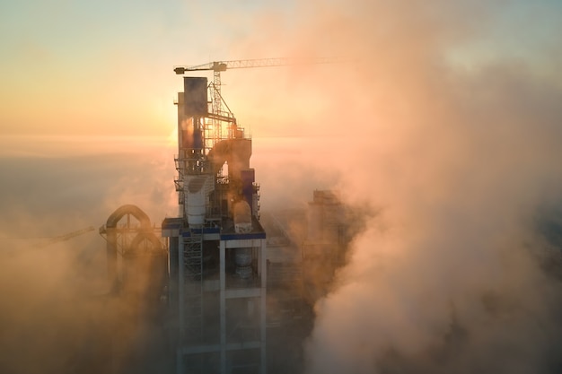 Aerial view of cement factory with high concrete plant structure and tower crane at industrial production site on foggy morning. Manufacture and global industry concept.