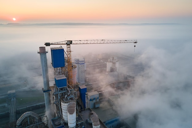 Aerial view of cement factory with high concrete plant structure and tower crane at industrial production site on foggy morning. Manufacture and global industry concept.