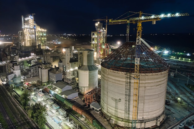 Aerial view of cement factory with high concrete plant structure and tower cranes at industrial production area at night. Manufacture and global industry concept.