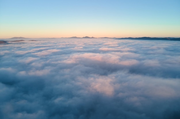 Aerial view of colorful sunrise over white dense fog with distant dark silhouettes of mountain hills on horizon