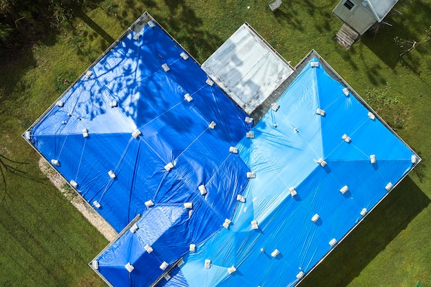 Aerial view of damaged in hurricane ian house roof covered with blue protective tarp against rain water leaking until replacement of asphalt shingles