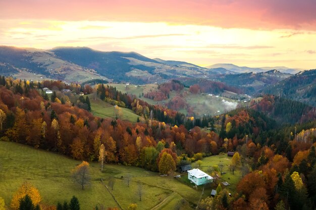 Aerial view of distant village with small shepherd houses on wide hill meadows between autumn forest trees in Ukrainian Carpathian mountains at sunset.