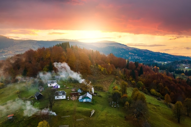 Aerial view of distant village with small shepherd houses on wide hill meadows between autumn forest trees in Ukrainian Carpathian mountains at sunset.