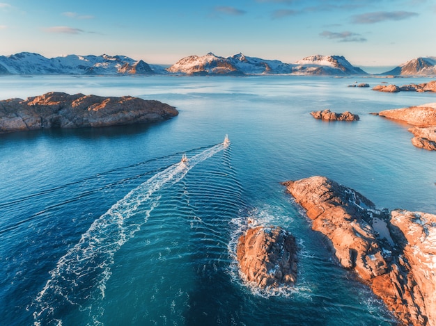 Aerial view of fishing boats, rocks in the blue sea, snowy mountains and colorful sky with clouds at sunset