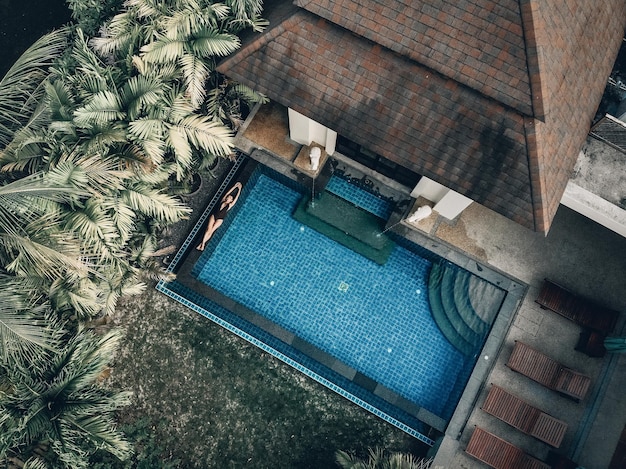 Aerial view of the hotel territory with palms, green lawn, tiled roof and the clear pool with a slim girl floating on the back; luxury concept.