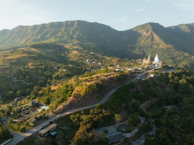 Una vista aerea di un paesaggio montuoso all'alba che mostra un sereno complesso di templi con una prominente statua bianca di buddha annidata tra la lussureggiante vegetazione