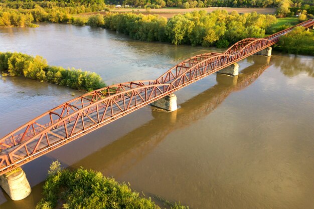 Aerial view of a narrow road bridge stretching over muddy wide river in green rural area.