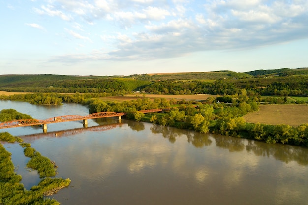 Aerial view of a narrow road bridge stretching over muddy wide river in green rural area.