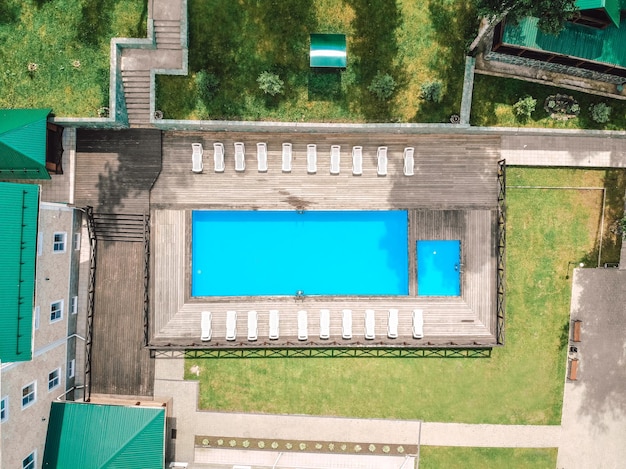 Вид с воздуха на открытый бассейн возле отеля и множество шезлонгов вокруг; зеленый газон, солнечный день; концепция отдыха.