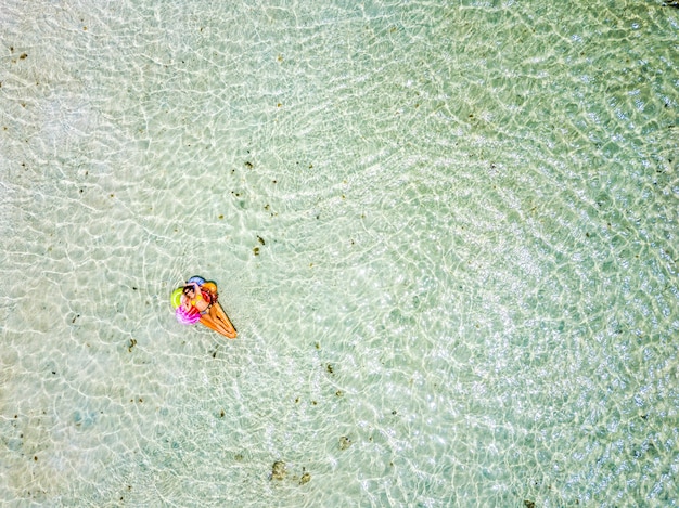 Aerial view of people in summer holiday vacation with beautiful girl on coloured trendy lilo relaxing and sunbathing on clear green ocean lagoon beach water