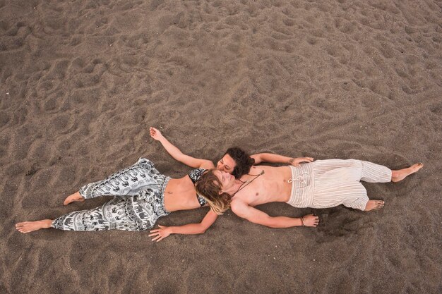 Aerial view picture of young cheerful beautiful couple laying down at the beach on the sand relax and enjoy the summer season in holiday ethnic and hippy clothes for nice people in vacation