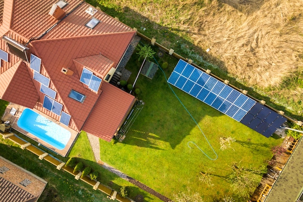 緑の芝生で覆われた庭、屋根の上のソーラーパネル、プール、風力タービン発電機のある民家の空撮。