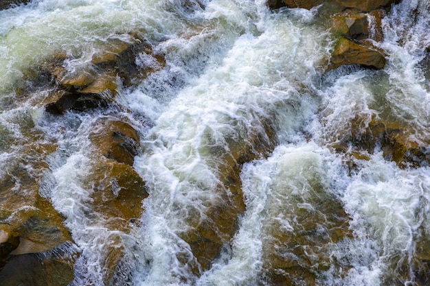 Вид с воздуха на речной водопад с чистой бирюзовой водой, падающей между мокрыми валунами с густой белой пеной.