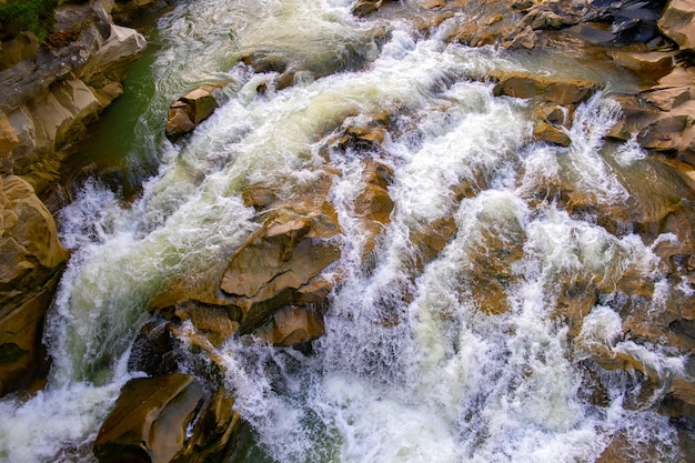 Вид с воздуха на речной водопад с чистой бирюзовой водой, падающей между мокрыми валунами с густой белой пеной.