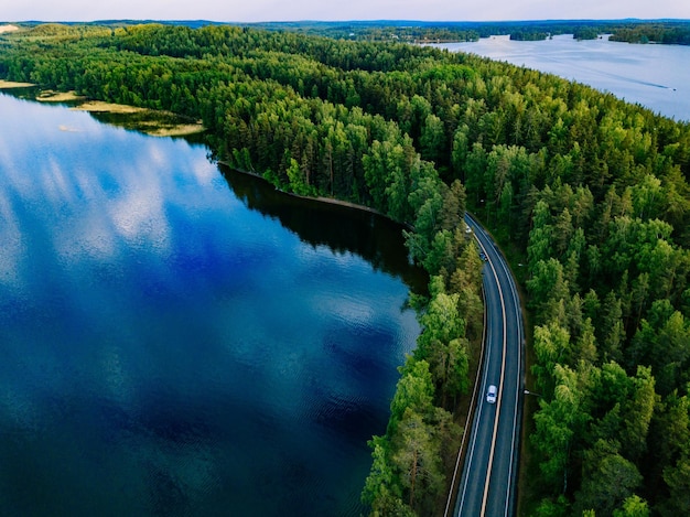 Aerial view of road with cars between green forest and blue lake water in summer Finland