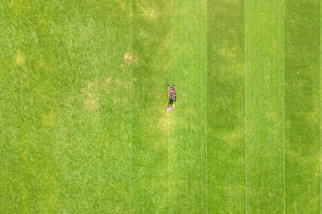 Aerial view of small figure of man worker trimming green grass with mowing mashine on football stadium field in summer.