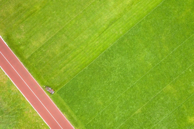 Aerial view of small figure of worker cutting green grass with mowing mashine on football stadium field with red running tracks in summer.