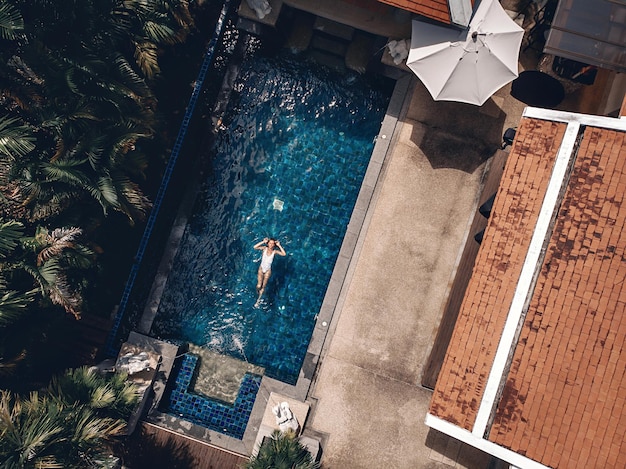 Aerial view of the territory of the Thai hotel: tiled terracotta roofs, palm trees, the pool with clear blue water and a young woman swimming there; drone.