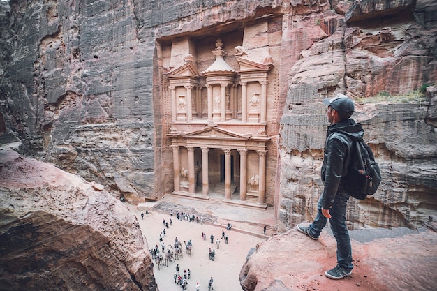 Aerial view of the Treasury with a hiker, solo traveler, young man tourist on a cliff after reaching the top, Al Khazneh in the ancient city of Petra, Jordan, UNESCO World Heritage Site