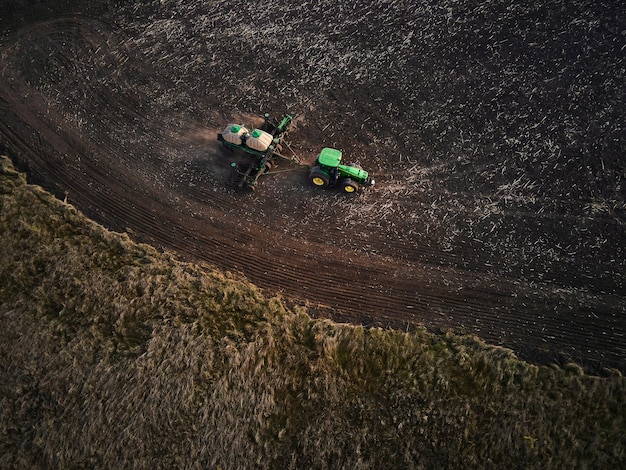 Aerial view of a two modern green tractors plowing dry agricultural field and seeding preparing land for sowing spring