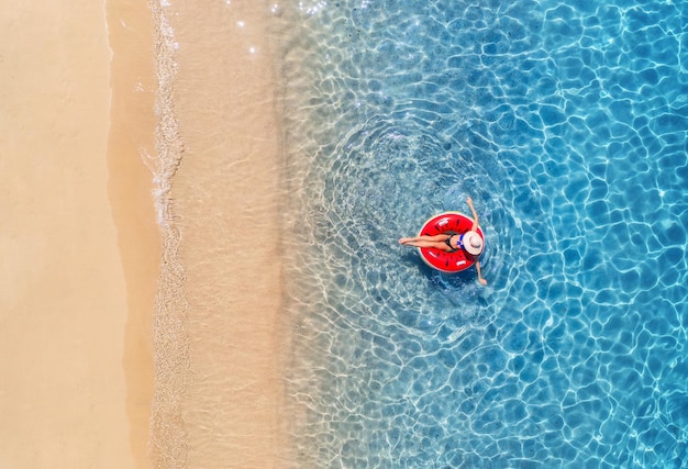 Aerial view of a woman in hat swimming with red swim ring in blue sea at sunrise in summer Tropical landscape with girl clear water waves sandy beach Top view Vacation Sardinia island Italy