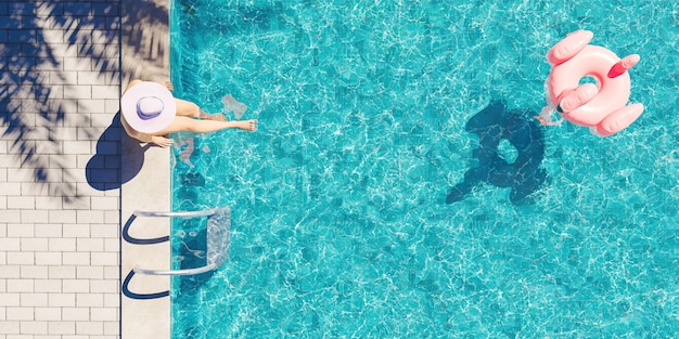 Вид с воздуха на женщину в шляпе, сидящую на краю бассейна с тенью пальмы и плавающим в воде фламинго