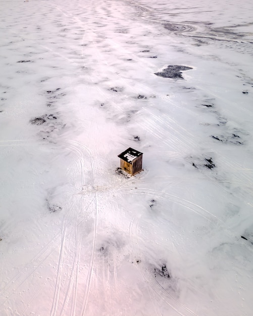 Aerial view of a wooden small hut in the middle of a frozen snow-covered field with ski tracks