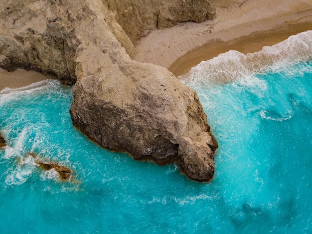 Aerial views of the turquoise sea water with wild seashore and waves reaching sandy beach and steeply rocks on a sunny day.