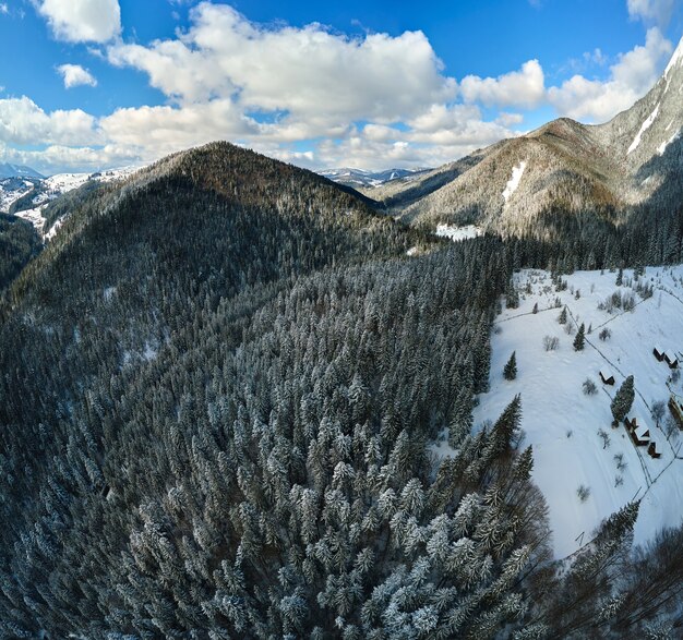 Aerial winter landscape with small rural houses between snow covered forest in cold mountains.