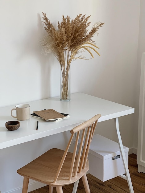 Aesthetic minimal office workspace interior design chair and\
table mug notebook pampas grass floral bouquet on white table\
against white wall girl woman boss work at home business\
concept