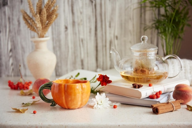本の上のティーポットとカボチャの形をしたお茶のカップに入った審美的なオーガニックリンデンティー 乾燥した香りのよい花 小麦 秋のアレンジメント イチジク 桃 家でリラックスできる居心地の良い雰囲気