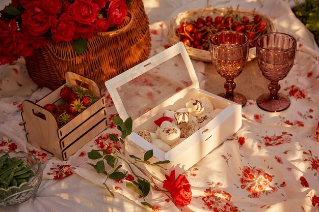 Aesthetic summer picnic in the sunny garden Pink tablecloth box of marshmallows strawberries cherry peas and glasses of wine among pink roses Romantic evening appreciation of the moment