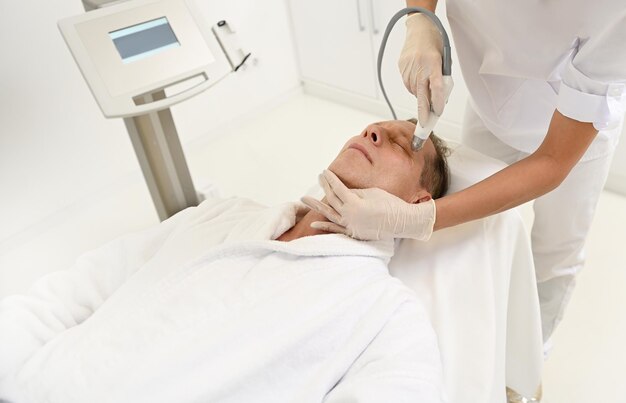 Aesthetician performs facelift procedure for mature man using modern ultrasonic equipment in SPA for exfoliation, stimulation and hydration. Men's aesthetic cosmetology and anti-aging skincare concept