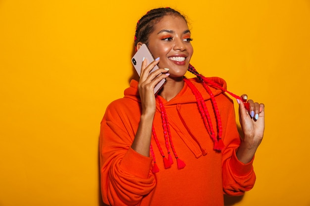 Afbeelding close-up van een vrolijke Afro-Amerikaanse vrouw die een hoodie-shirt draagt en glimlacht terwijl ze op een mobiele telefoon praat, geïsoleerd over geel