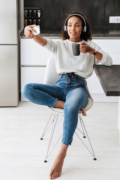 Afbeelding van aantrekkelijke Afro-Amerikaanse meisje hoofdtelefoon nemen selfie foto, zittend in lichte keuken