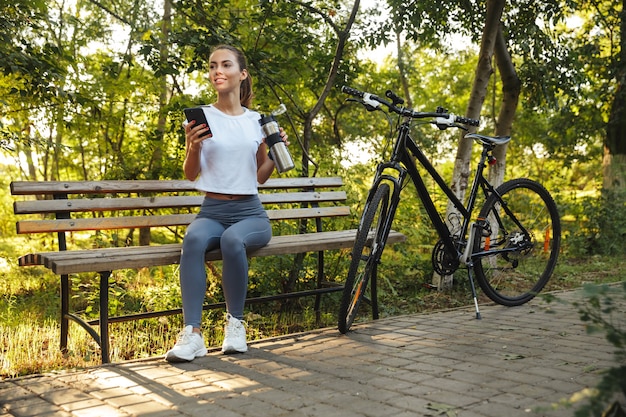 Afbeelding van aantrekkelijke vrouw zittend op een bankje in park met fiets, en met behulp van mobiele telefoon