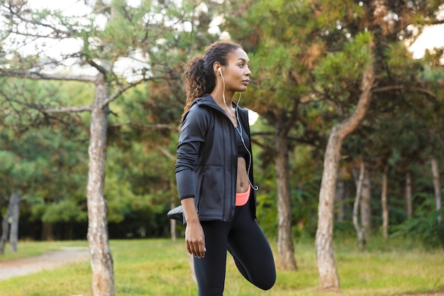 Afbeelding van Afro-Amerikaanse vrouw 20s dragen zwarte trainingspak uit te werken, en haar lichaam uitrekken in groen park