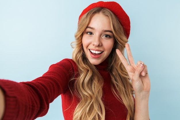 Afbeelding van bescheiden blonde vrouw 20s met rode baret glimlachen tijdens het nemen van selfie foto, geïsoleerd