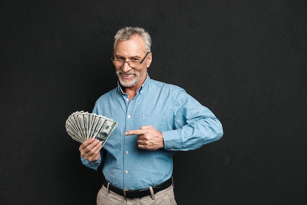 Afbeelding van blanke man van middelbare leeftijd 60s met grijze haren wijzende vinger op geldprijs met veel dollar biljetten, geïsoleerd over zwarte muur