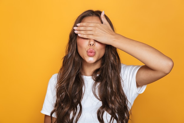 Afbeelding van blanke mooie vrouw 20s met lang haar dat ogen bedekt met handen, geïsoleerd over gele muur