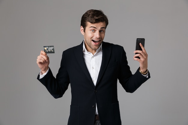 Afbeelding van brunette man 30s in pak met zwarte mobiele telefoon en creditcard, geïsoleerd over grijze muur