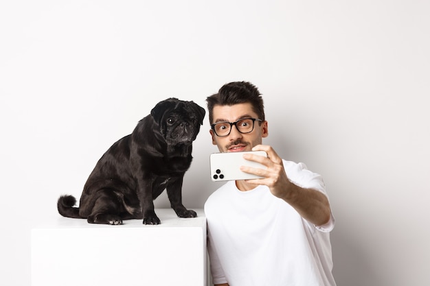 Afbeelding van de knappe jongeman die selfie met schattige zwarte hond op smartphone, poseren met pug over wit