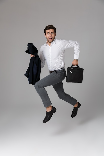 Afbeelding van de volledige lengte van jonge man 30s in pak met tas en jas in handen, geïsoleerd over grijze muur
