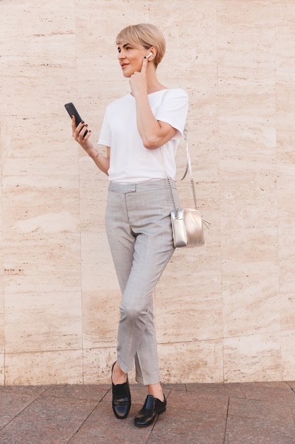 Afbeelding van de volledige lengte van stijlvolle blonde vrouw, gekleed in een wit t-shirt met behulp van mobiele telefoon, terwijl staande tegen een beige muur buiten en bluetooth oortelefoon aan te raken