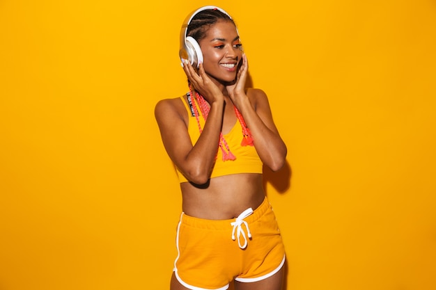 Afbeelding van een aantrekkelijke Afro-Amerikaanse vrouw die afro-vlechten draagt, glimlachend en luisterend naar muziek met een koptelefoon geïsoleerd over een gele muur