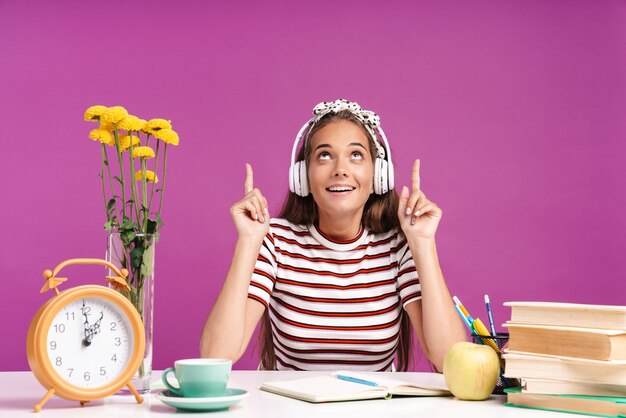 Afbeelding van een aardig lachend meisje dat met de vingers naar boven wijst en een koptelefoon gebruikt terwijl ze aan een bureau zit geïsoleerd over een violette muur