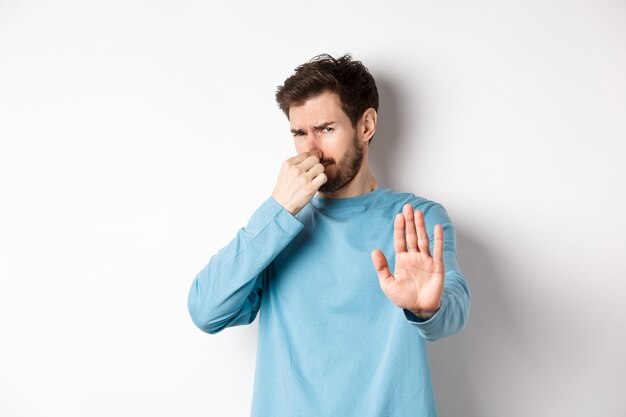 Afbeelding van een blanke man strekt zijn handen uit om iets te stoppen of af te wijzen, sluit neus walging, weigert product met slechte geur, staande op witte achtergrond