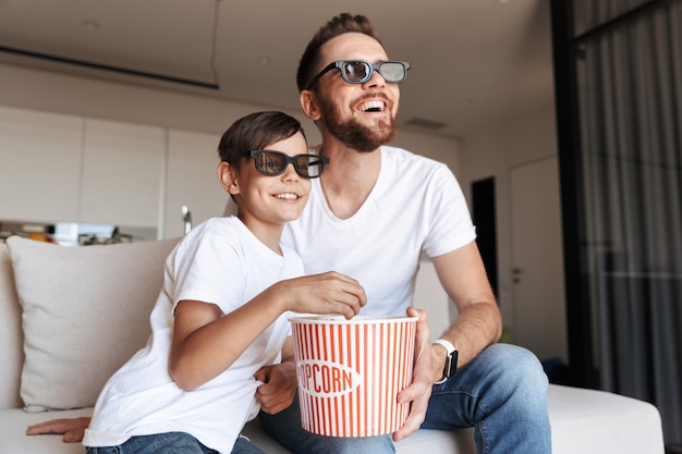 Afbeelding van een blanke vader en zoon die een 3D-bril draagt die popcorn eet en lacht, zittend op de bank thuis en film kijken