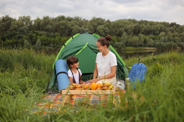Afbeelding van een blije mooie moeder die picknickt in de buurt van de rivier met haar dochter zittend op een plaid op de grond in de buurt van een tent, pratend en genietend van hun camping